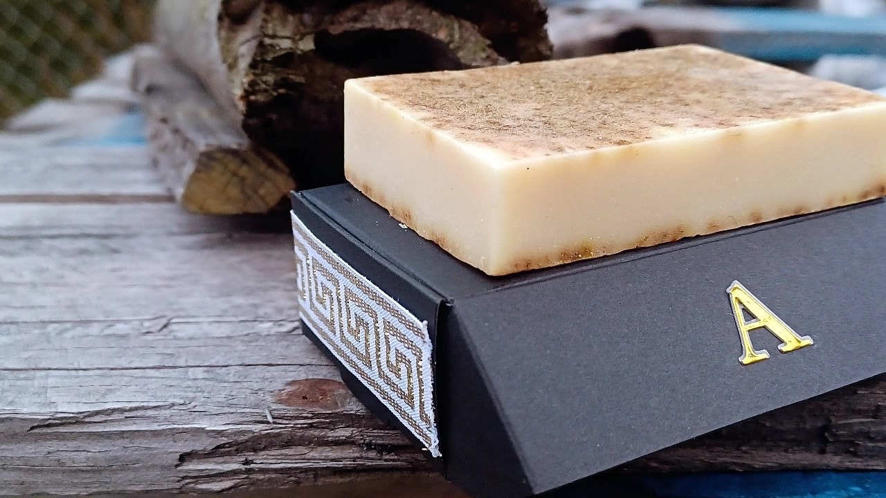 An APSTERGO soap bar resting on top of its matte-black box with a gold A and Greek key band, photographed outdoors on weathered wood